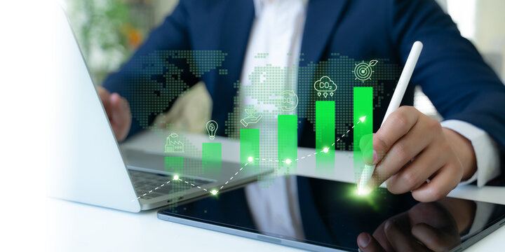 Carbon emission reduction target, green economy and sustainability concept. Business professional analyzes digital data visualizations on a tablet, featuring rising green bars and carbon-related icons