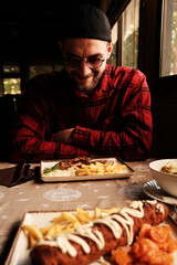 A man in a red plaid shirt sits at a table in a cozy restaurant with traditional Balkan food in front of him. Warm natural light enters from the window.