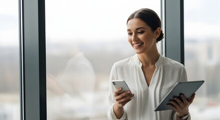 Smiling businesswoman uses tablet and smartphone near large window.