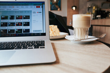 Laptop is open on a table with a glass of coffee and a plate of cake