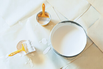 Open can of white paint with two paintbrushes and scattered drips on paper shows preparation and energy of a creative renovation moment.