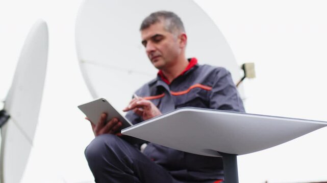 A male engineer installs and adjusts a satellite dish on the roof of an office building. 