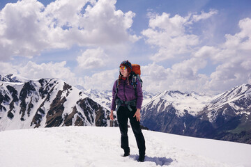 A tourist girl with a large backpack stands on a mountain pass in the snow with a view of the mountain peaks. Extreme travel