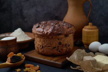 A round loaf of easter cake sits on a wooden board, surrounded by eggs, butter, flour, sugar, yeast and dried fruits