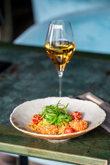 A glass of white wine and a bowl of pasta with shrimp, cherry tomatoes, and greens on a rustic table. The dish is garnished with sesame seeds and fresh herbs, creating an appetizing