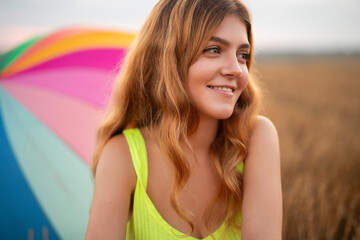A cheerful young woman enjoying an outdoor setting with a colorful umbrella behind her, standing...