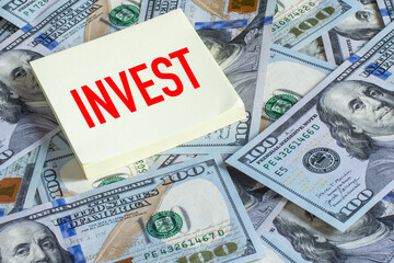 The word ‘INVEST’ written on yellow office sticker cardstock placed on hundred-dollar bills. Business and finance concept.