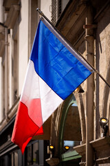 French flag. National colors: blue, white and red. Hanging on a flag pole in Paris (France). Tricolore “bleu blanc rouge“ blown by wind with blurred facade of an old houses and backlit by sunlight.