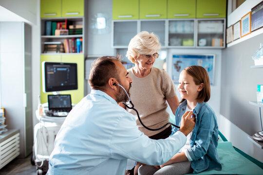 Doctor examining young girl with stethoscope while grandmother watches in medical clinic