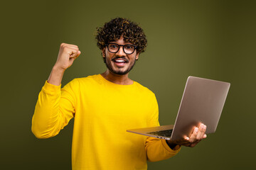 Young man with glasses happily seated holding a laptop while gesturing triumphantly on a khaki...