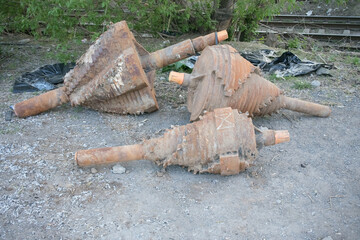Three large, weathered metal pieces resembling industrial drill rest on gravel. Surrounding vegetation and a hint of tracks suggest an abandoned site in daylight.