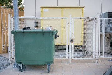 Green trash bin positioned near a fenced utility area behind a building in late afternoon light