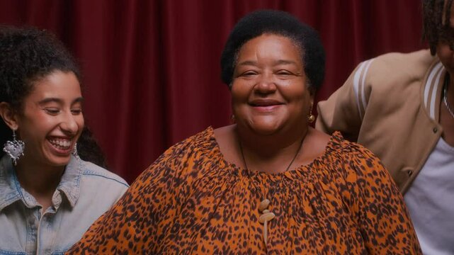 Chest up portrait of senior Black woman in leopard print blouse sitting calmly as her two smiling young adult grandkids leaning in with heads resting on her shoulders in cozy red-curtained photo booth