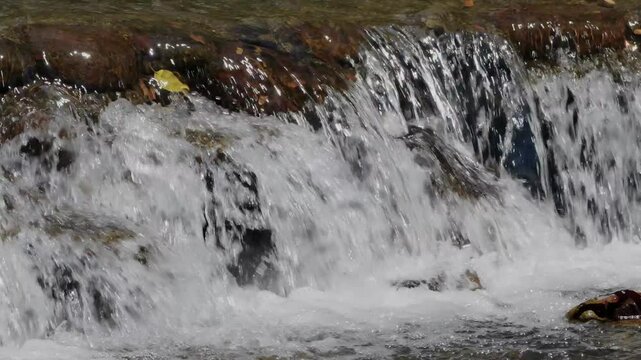 Several small cascades of river water falling through an elevated stone ledge in sunny weather cerating foams and splashes in slow motion.