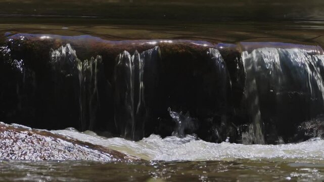 View of small cascades of river water falling through an elevated stone ledge in sunny weather cerating foams and splashes.