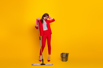Stylish young woman in a vibrant red suit posing with cleaning tools against a vivid yellow...