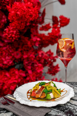 A gourmet dish with roasted meat, fresh vegetables, and artistic sauce on a white plate, accompanied by a citrus-infused beverage. The background features vibrant red flowers