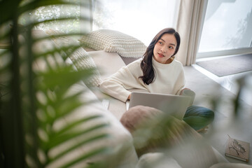 Young woman sitting on the floor with a laptop and coffee mug, relaxing in a cozy home with soft light and greenery.