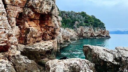Steep rocky cliffs with textured orange and white stone rise above calm turquoise water. Dense greenery crowns the distant headland under an overcast sky. Mediterranean Sea, Turkey, coastal, travel.