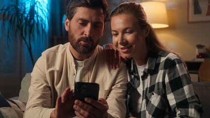 Caucasian family couple man woman home evening using smartphone together online shopping on sofa husband wife girl guy buying internet purchase choose goods home renovation night browsing mobile phone