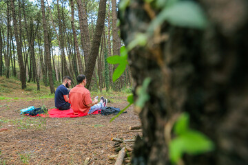 Two men in the forest resting and enjoying nature. Lying on a red blanket, reading, eating, and chatting