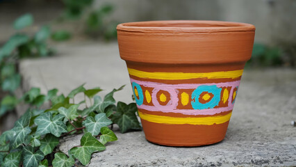 Colorful painted flower pot placed on a stone surface surrounded by green ivy leaves illustrating artistic creativity and nature’s beauty