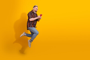Obraz premium Cheerful middle aged man holding a smartphone while jumping against yellow background showcasing energy and joy