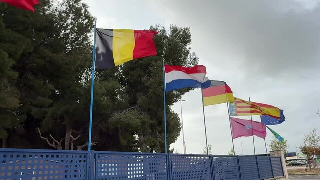 National flags of belgium, netherlands, catalonia, and eu waving on metal poles, positioned behind blue fencing with cloudy background