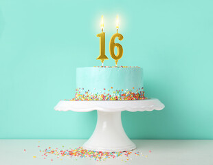 Yummy cake with burning number shaped candles and sprinkles on white table against turquoise background. 16th birthday or other special occasion