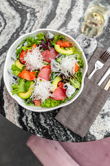 Fresh gourmet salad with mixed greens, avocado, salmon, tuna, grapefruit, and daikon radish in a white bowl on a marble table. Accompanied by white wine and cutlery, ideal for healthy dining