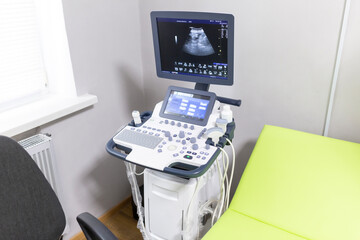 A modern ultrasound machine with a screen displaying a scan, an examination chair, and a patient table are in a hospital echo room.