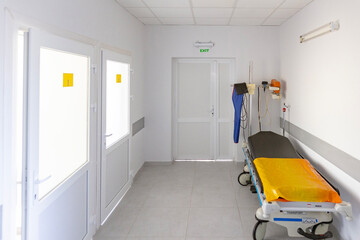 A sterile hospital admission department corridor features a modern stretcher, a mobile X-ray machine, and bright white doors, including an exit.