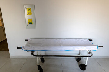A clean hospital stretcher with a white sheet and blue trim is positioned in a clean, white-walled medical facility hallway.