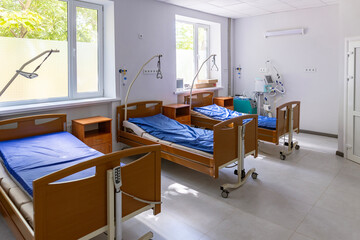 A multi-bed hospital ward features three empty beds, each with a lung ventilator, oxygen concentrator, and oxygen hub, in a brightly lit, sterile environment.