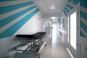 A hospital admission department corridor features striking blue and white diagonal stripes on the walls, with an empty stretcher and open doors leading further into the facility.