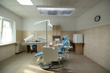 A modern dental clinic room features a blue dental chair, white dental unit with instruments, overhead operating light, and wooden cabinetry in a clean treatment space.