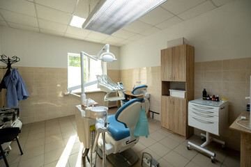 A modern dental clinic room features a blue dental chair, white dental unit with instruments, overhead operating light, and wooden cabinetry in a clean treatment space.