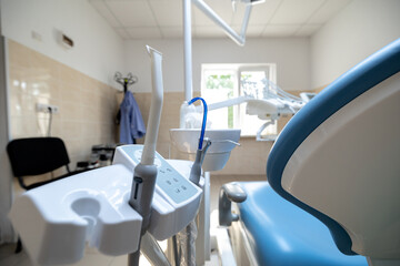 A modern dental clinic features a blue dental chair with associated instruments, including suction and handpieces, in a bright, clean treatment room.