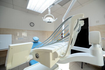 A modern dental clinic room features a blue dental chair, white dental unit with instruments, an overhead operating light, and a wall clock in a clean setting.