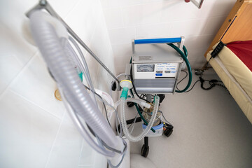 A ventilator, a complex medical apparatus with tubing and controls, stands next to a hospital bed in a clean, tiled room