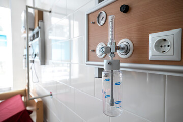 An oxygen hub with a flow meter and humidifier bottle is mounted on a wooden-paneled wall above a hospital bed in an intensive care unit