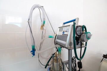 A ventilator, a complex medical apparatus with tubing and controls, stands next to a hospital bed in a clean, tiled room