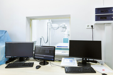 A control room for an X-ray diagnostic system features multiple computer monitors and keyboards, overlooking the examination room through a large window.