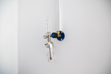 An oxygen flow meter with a humidifier bottle is mounted on a white wall, part of an oxygen hub, ready for use in a hospital intensive care unit.