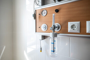 An oxygen hub with a flow meter and humidifier bottle is mounted on a wooden-paneled wall above a hospital bed in an intensive care unit