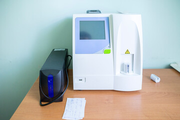 A white medical blood analyzer with a display and a black uninterruptible power supply sit on a wooden table in a clinic room.