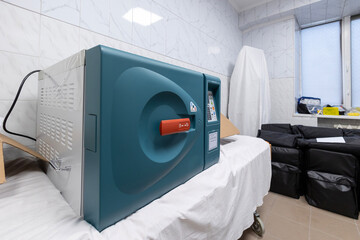 A new, unplugged medical sterilizer, an autoclave, rests on a gurney in a clean hospital room with tiled walls and nearby equipment bags.