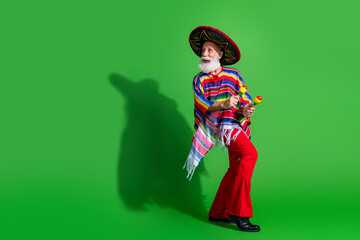 Cheerful elderly man in festive traditional Mexican attire holding maracas on a bright green background