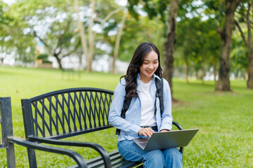 Asian female student using laptop and smiling while sitting on bench in park, enjoying outdoor learning environment and taking advantage of wireless technology