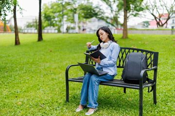 Asian female student sitting on a bench in a park studying with a laptop and a notebook taking notes, enjoying the fresh air and the green environment